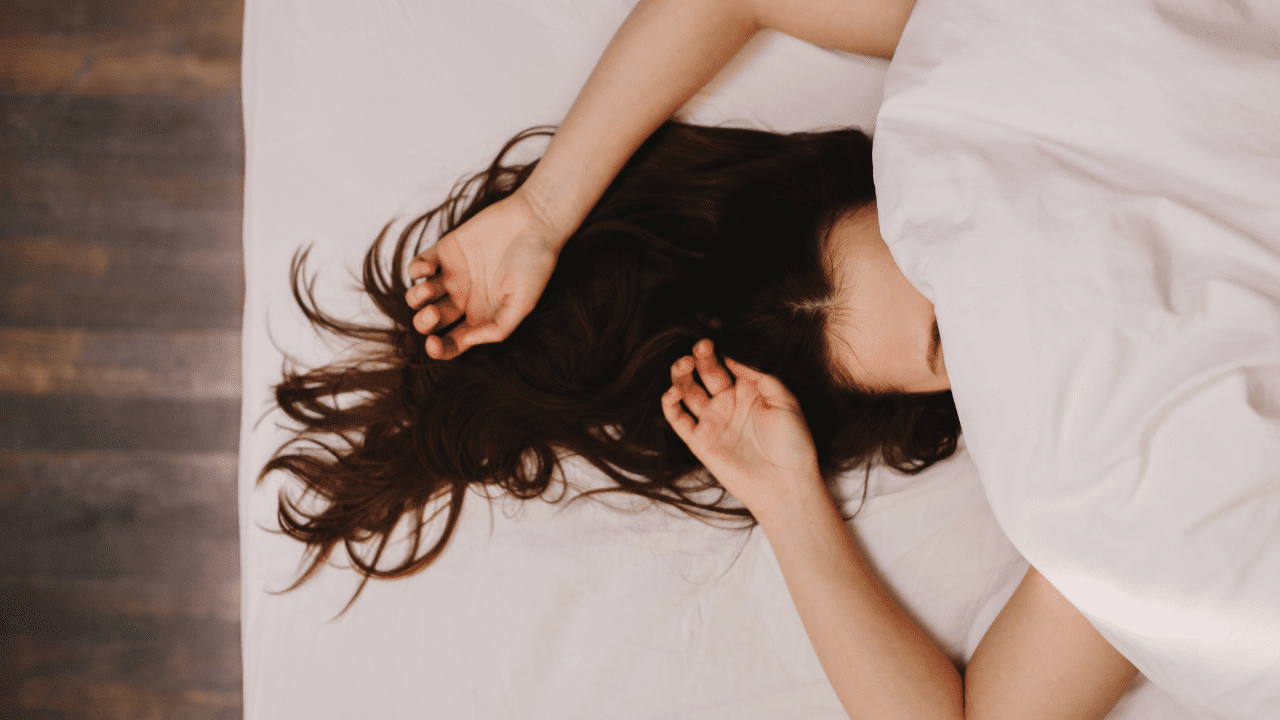 A girl lays in bed with her face covered by a blanket and her hands over her head.
